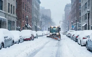 Verschneite Autos parken am Straßenrand während ein Schneepflug die Straße räumt.